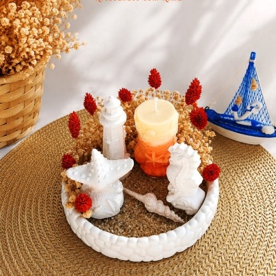 Base decorativa com vela, figuras marinhas brancas e flores secas em tapete ráfia com cesto de flores e barco azul ao lado.