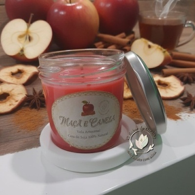 Vela artesanal de maçã e canela em frasco de vidro com tampa metálica, rodeada por maçãs, canela e chá quente.