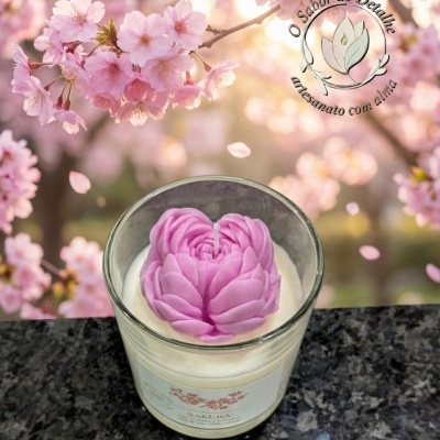 Vela em copo de vidro com topo em forma de flor cor-de-rosa sobre superfície preta e flores de cerejeira ao fundo.