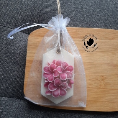 Vela hexagonal branca com flores rosa em saco de organza numa base de madeira com texto e logotipo pretos.