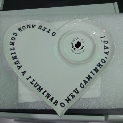 Prato cerâmico branco em forma de coração com texto preto e selo no centro em fundo branco.