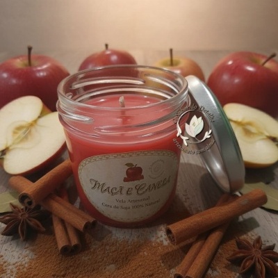 Vela artesanal vermelha em frasco de vidro com rótulo maçã e canela, rodeada por paus de canela e maçãs em fundo de madeira.
