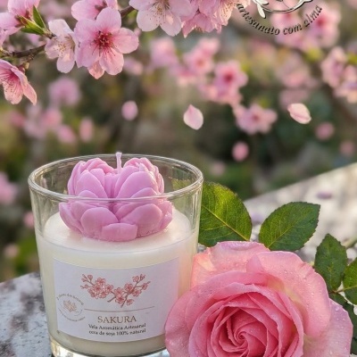 Vela artesanal com topo em flor cor-de-rosa e rosa natural em superfície escura com flores de cerejeira ao fundo