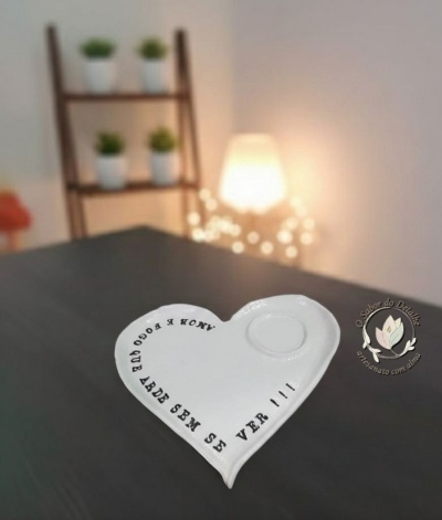 Prato cerâmico branco em forma de coração com texto preto e relevo, em superfície preta, com estante e luz difusa ao fundo.