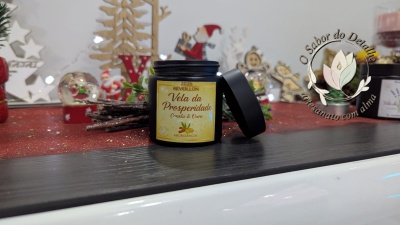 Vela da Prosperidade em pote preto com rótulo amarelo e decoração natalícia em fundo