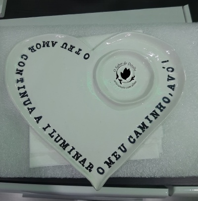 Prato cerâmico branco em forma de coração com texto preto e selo no centro em fundo branco.