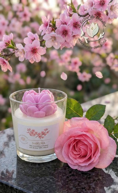 Vela artesanal com topo em flor cor-de-rosa e rosa natural em superfície escura com flores de cerejeira ao fundo