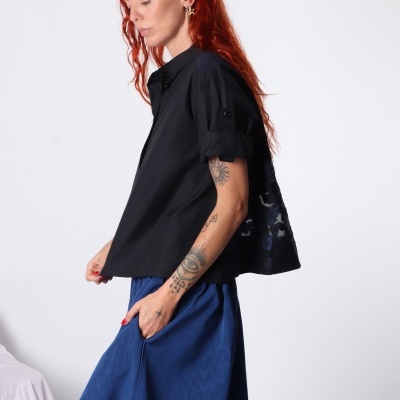 Mulher com camisa preta de manga curta e saia azul comprida, com tatuagens no braço.