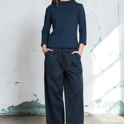 Mulher com blusa azul, calças largas azul e sandálias verdes em fundo de parede branca