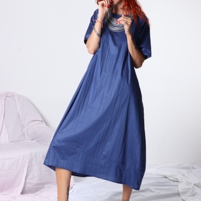Mulher com vestido azul e sandálias azuis a posar em fundo branco com tecidos brancos e rosa no chão
