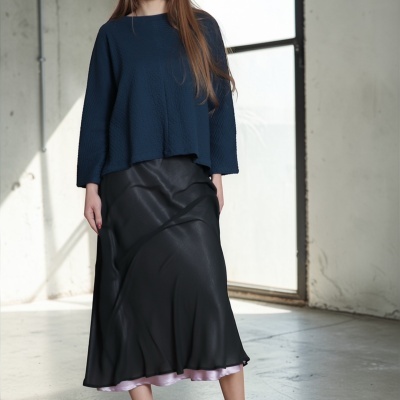 Mulher de blusa azul, saia preta com detalhe lilás e sandálias verdes em ambiente interno