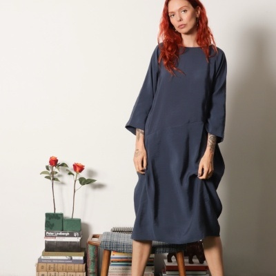 Mulher com vestido azul escuro e sandálias castanhas em ambiente com tapete verde e livros