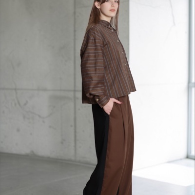 Mulher com camisa castanha às riscas e calças largas castanhas com faixa preta, calçado bege, em espaço interior cinzento.