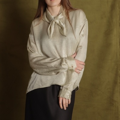 Camisa bege de cetim com nó no colarinho e botões nas mangas, usada com saia preta em fundo verde-oliva.