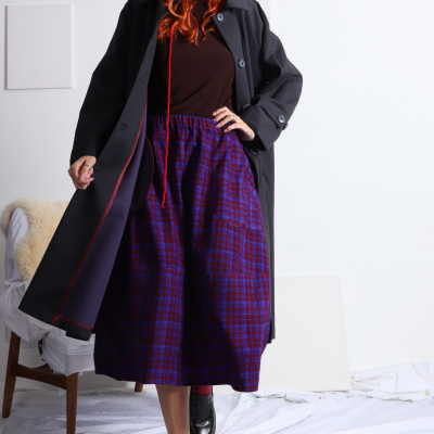Mulher de pé com vestido xadrez azul e vermelho, casaco preto, sapatos pretos e meias vermelhas, em estúdio com fundo branco.