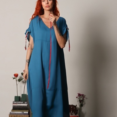 Mulher ruiva com vestido azul e mangas com pregas, em cenário interior com livros e flores.