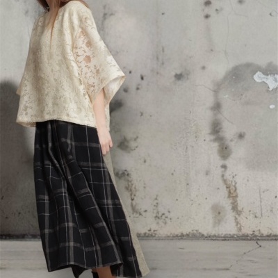 Mulher com blusa creme floral e saia xadrez preta em fundo de parede cinza.