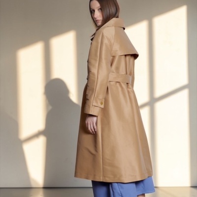 Mulher com casaco camel longo, culotes azul claro e sandálias azuis em fundo neutro com sombra de janela