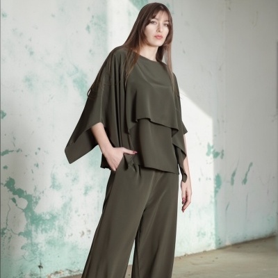 Conjunto de roupa feminina verde-oliva com blusa de manga larga e calças largas, com ténis cinzentos, em ambiente interior com parede envelhecida.