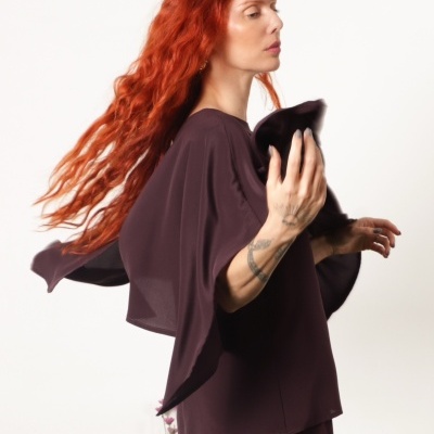 Mulher com cabelo ruivo usando roupa castanha escura com mangas largas e fundo branco