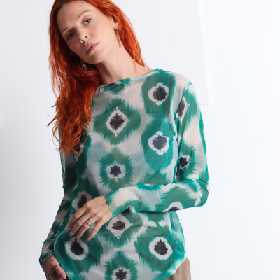 Mulher a usar blusa verde com padrão e calças bege em fundo branco