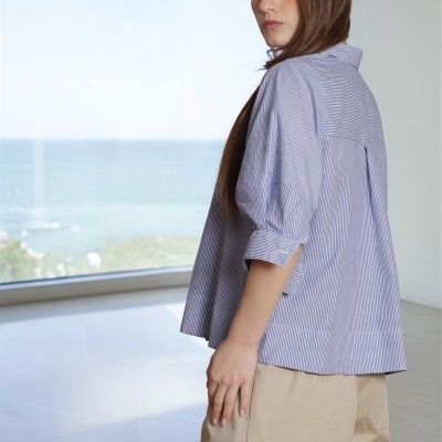 Mulher de perfil a usar blusa às riscas azul claro e branco com calças bege, em interior com vista para o mar