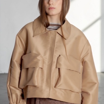 Casaco bege claro acetinado com bolsos grandes, mulher com saia xadrez castanha e blusa riscada.