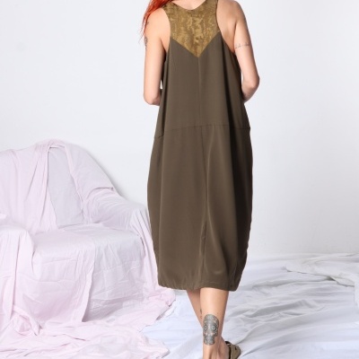 Vestido feminino verde-oliva de costas com detalhe dourado, cabelo ruivo, sandálias douradas, cadeira rosa ao fundo