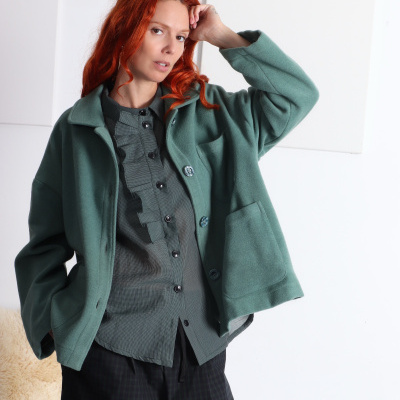 Mulher com casaco verde de lã, camisa cinza com folhos e calças pretas xadrez em fundo branco