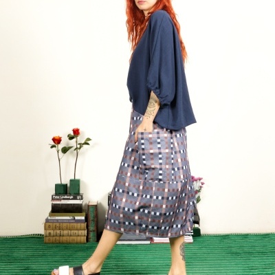 Mulher de perfil com blusa azul, saia xadrez e sandálias em tapete verde.