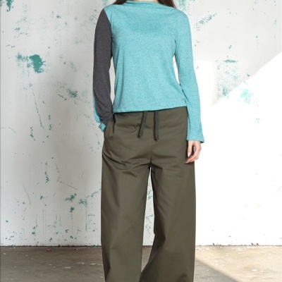 Mulher com camisola azul clara, calças largas caqui e sapatos cinzentos em fundo de parede branca com manchas verdes
