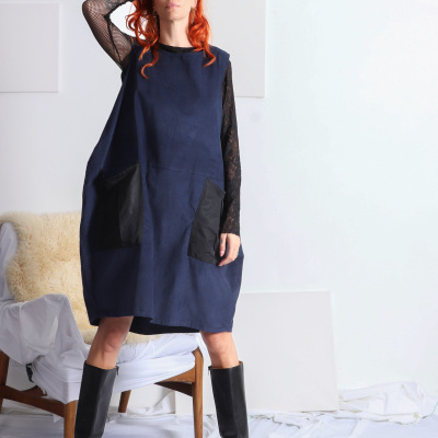 Mulher com vestido azul escuro e bolsos pretos, camisa preta de renda e botas pretas, em ambiente minimalista