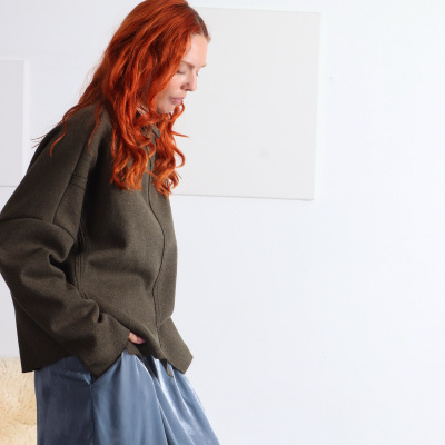 Mulher ruiva a usar camisola verde-militar e saia azul-cinzenta contra fundo branco.