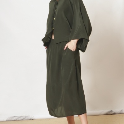 Modelo veste conjunto verde-oliva com camisa e saia, usando sandálias preto e branco em fundo branco