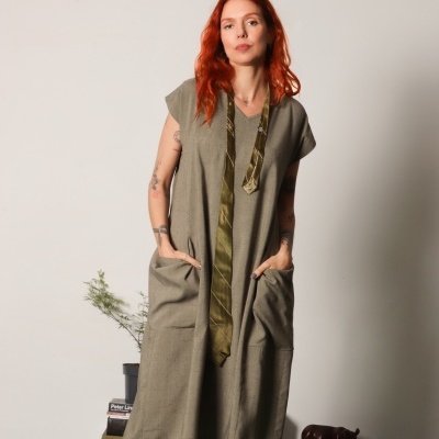 Mulher com vestido verde-oliva de tecido leve com bolsos e mangas curtas, em ambiente interior com livros e planta ao fundo