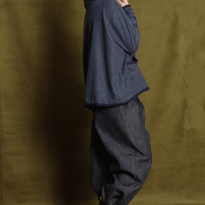 Pessoa com casaco azul e calças largas cinza e botas castanhas em fundo verde-oliva