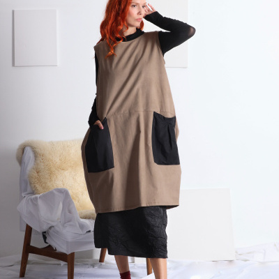 Mulher vestindo vestido bege com bolsos pretos, blusa preta, sapatos pretos e meias vermelhas em ambiente branco com cadeira de madeira