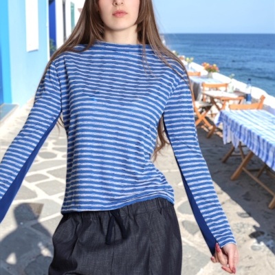 Camisa de riscas azuis e cinzentas com calças largas pretas usada por pessoa jovem junto ao mar com mesas e cadeiras de madeira.