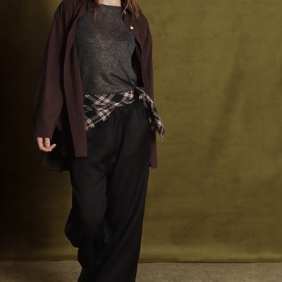 Modelo com camisa castanha aberta, camisola cinza escura, calças pretas largas, botas pretas e camisa xadrez amarrada à cintura.