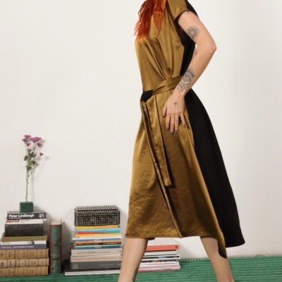 Vestido midi dourado e preto acetinado com cinto, sandálias pretas, mulher com tatuagens e cabelo ruivo, livros empilhados e flores em jarro.