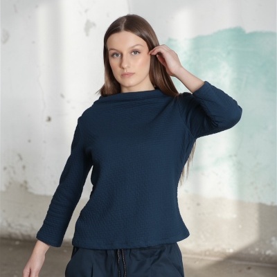 Mulher com blusa azul escura texturizada e calças largas azuis num espaço interior