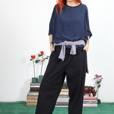 Mulher ruiva com roupa azul e preta e cinto listrado, sandálias, em tapete verde com livros e flores ao fundo