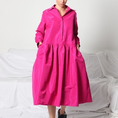 Mulher a usar vestido comprido rosa vibrante com gola e bolsos, a caminhar sobre chão branco.