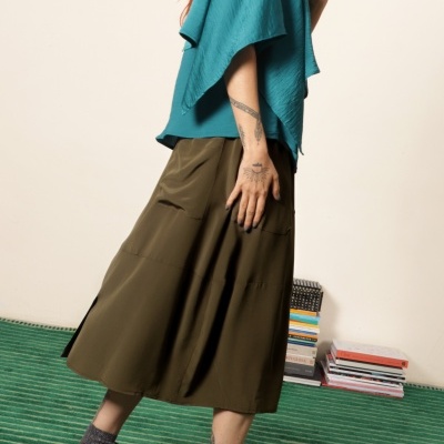 Pessoa usa blusa azul turquesa, saia verde militar, sapatilhas desportivas azuis e meias cinzentas com brilho em sala com tapete verde e livros.