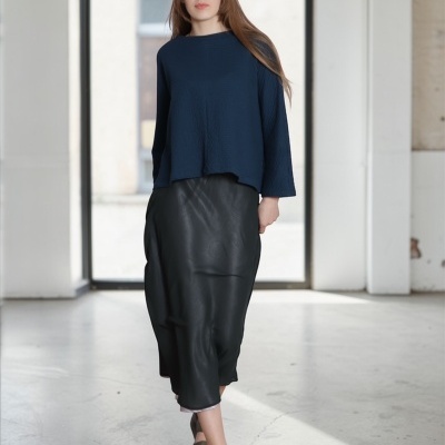 Mulher vestindo camisola azul, saia preta tipo couro e sandálias verdes num espaço interior