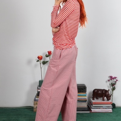 Mulher com roupa às riscas vermelhas e brancas e calças largas vermelhas com padrão xadrez, sandálias azuis, em casa com tapete verde e decoração de livros e flores.