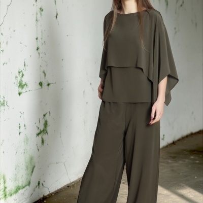 Mulher com conjunto verde-oliva em tecido fluido, blusa com sobreposição e calças largas, sapatos cinzentos em fundo de parede branca rachada