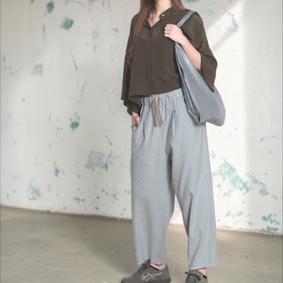 Mulher com camisa verde-oliva, calças largas cinza claro e ténis cinzentos, com bolsa cinza claro, em ambiente com parede desgastada.