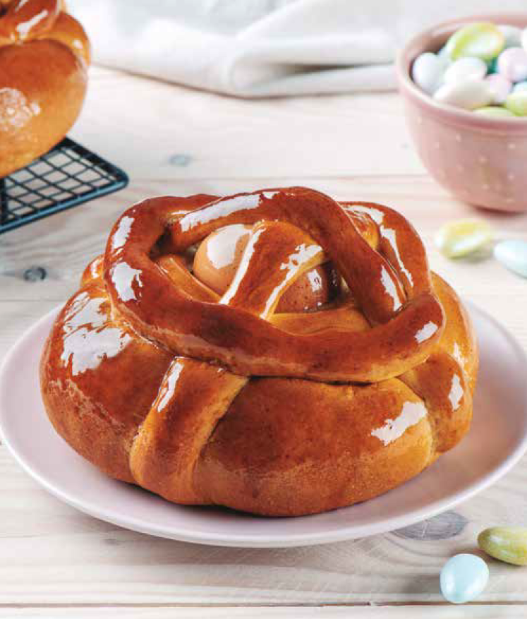 Credi Folar (500gr. / 5 Kilos) und. Pão doce com ovo cozido e brilho, em prato branco, com tigela de amêndoas coloridas ao fundo.