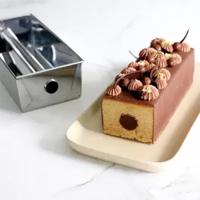 Bolo retangular com cobertura e decoração de chocolate numa travessa branca ao lado de uma forma metálica